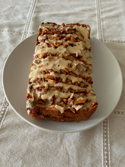 A loaf of Vermont Maple Apple Fritter Bread drizzled with a creamy glaze on a white plate sitting on top of a white table cloth.