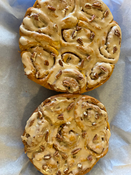 Two rounds or pumpkin cinnamon buns topped with cream cheese frosting and pecans.