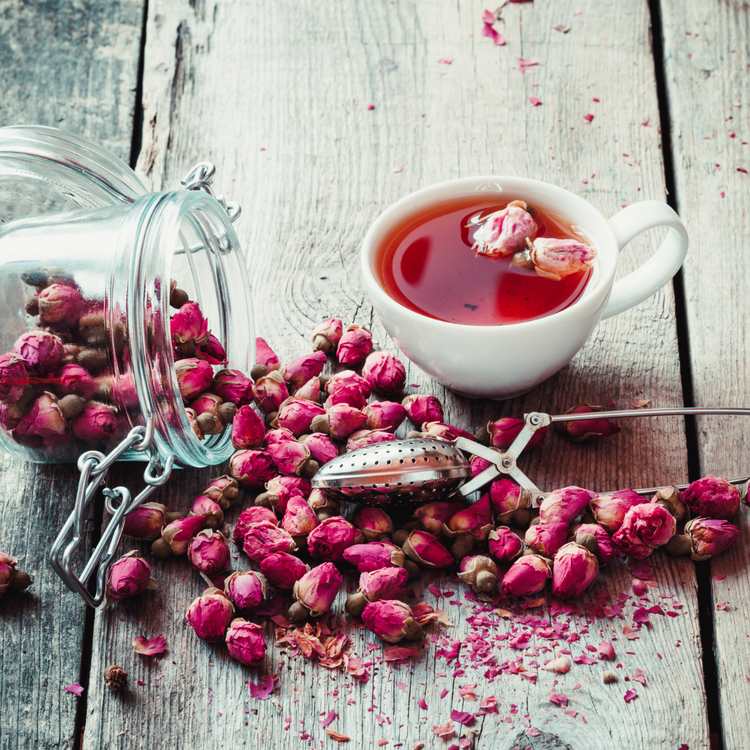 A white cup of rosehip tea with a spoon next to it and a jar with rosehips spilled out across a gray wooden table surrounding the tea cup.